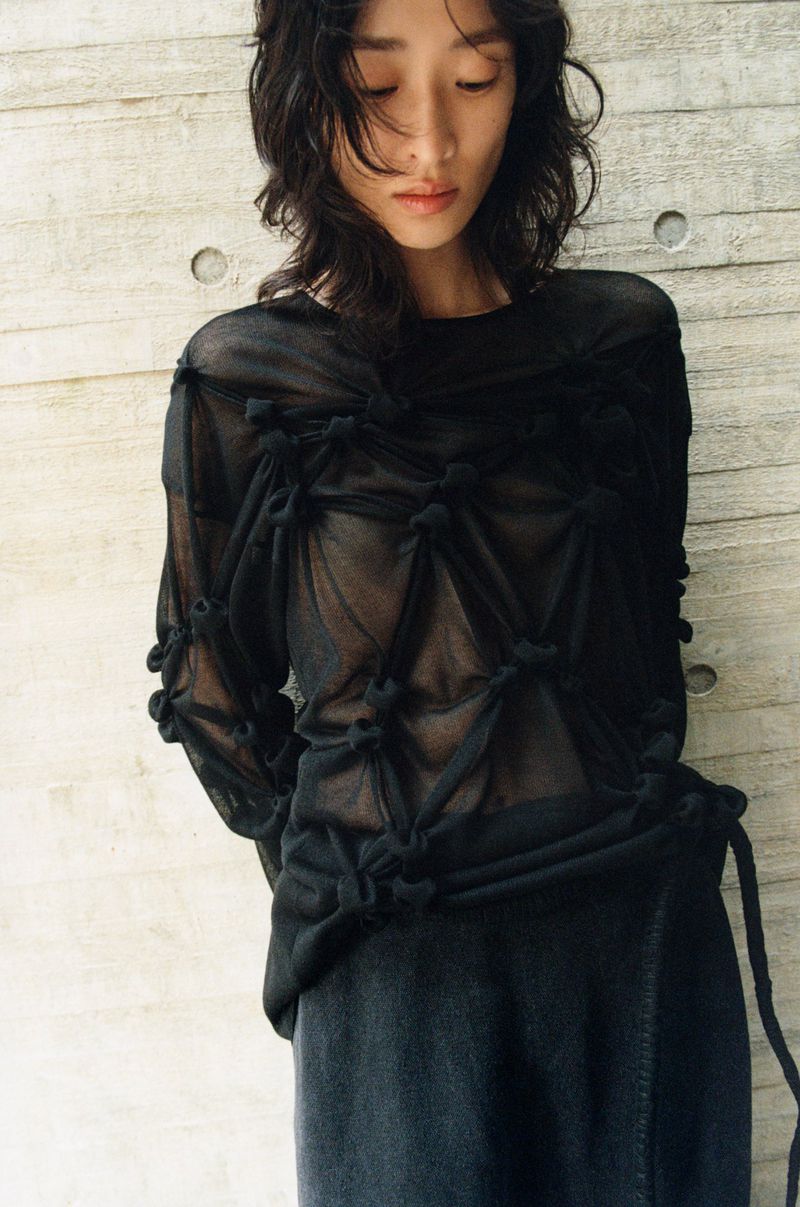 Indoor shot of an Asian fashion model wearing a black sheer knotted long-sleeved top with a black skirt against a textured gray wall.