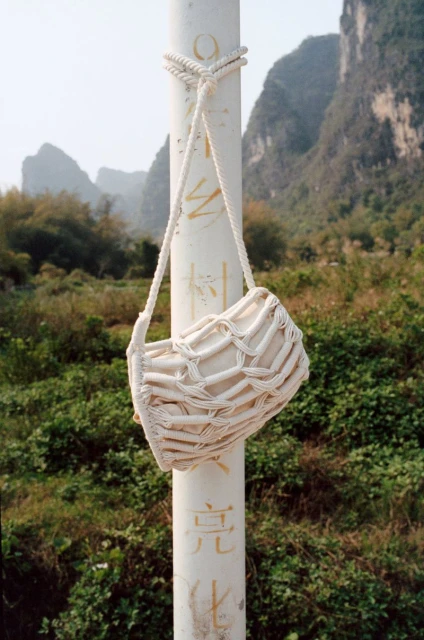 A cream-colored woven shoulder bag hangs from a white pole, with a landscape of green vegetation and mountains in the background.