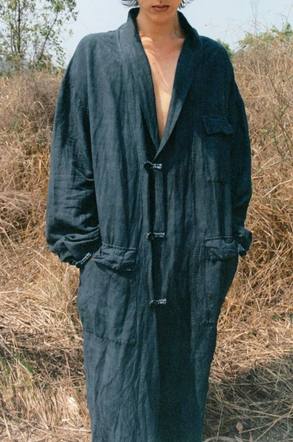 Person in a long, dark blue robe poses in a field of dry grass. The individual has an unbuttoned collar and short dark hair.