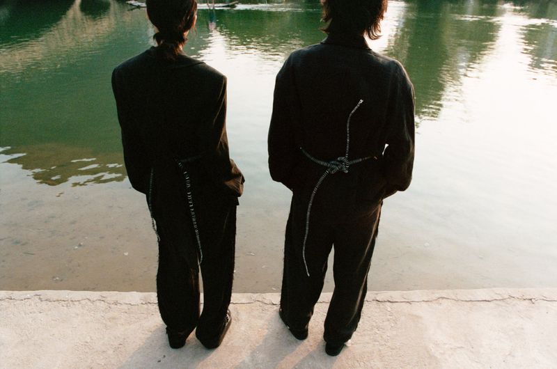 Two slender adults stand near a pond with sunlight reflecting on the water, backs to the camera, wearing dark matching pantsuits.