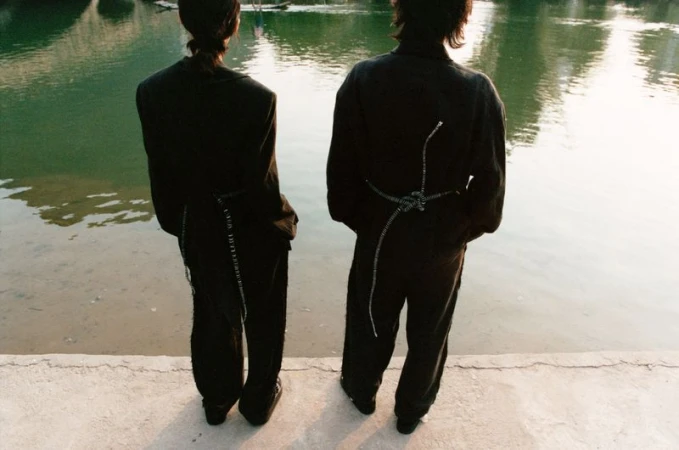Two slender adults stand near a pond with sunlight reflecting on the water, backs to the camera, wearing dark matching pantsuits.