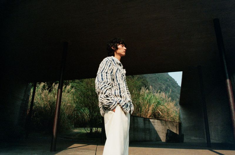 An adult male poses in an outdoor setting. The man is wearing a striped shirt and cream pants. The background shows green foliage.