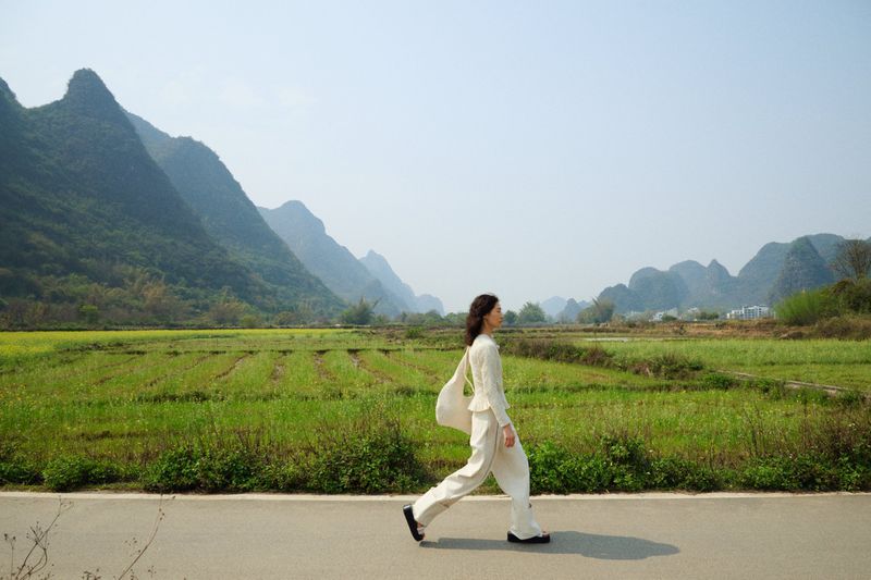 Woman in white suit walks on path with mountain backdrop. Fashion editorial shot in nature, side view, full body, green fields.