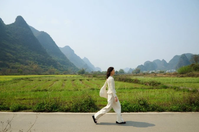 Woman in white suit walks on path with mountain backdrop. Fashion editorial shot in nature, side view, full body, green fields.