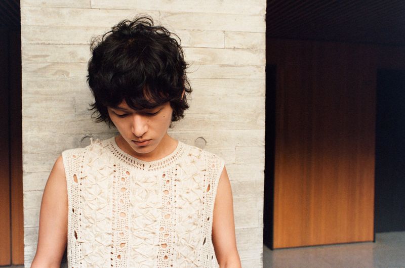 Young adult poses indoors wearing a cream-colored knit tank top; neutral background with natural light and concrete wall.