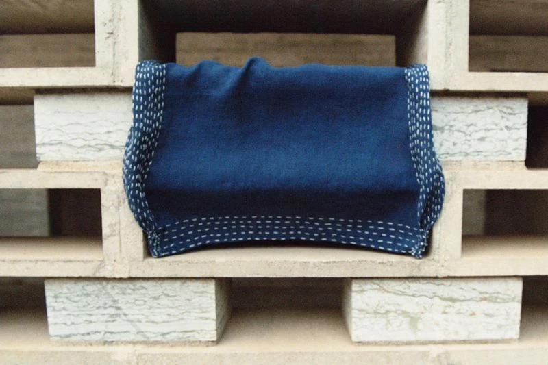 Close-up of a folded indigo-dyed textile with white stitching, resting on a neutral-toned concrete shelf with a geometric pattern.
