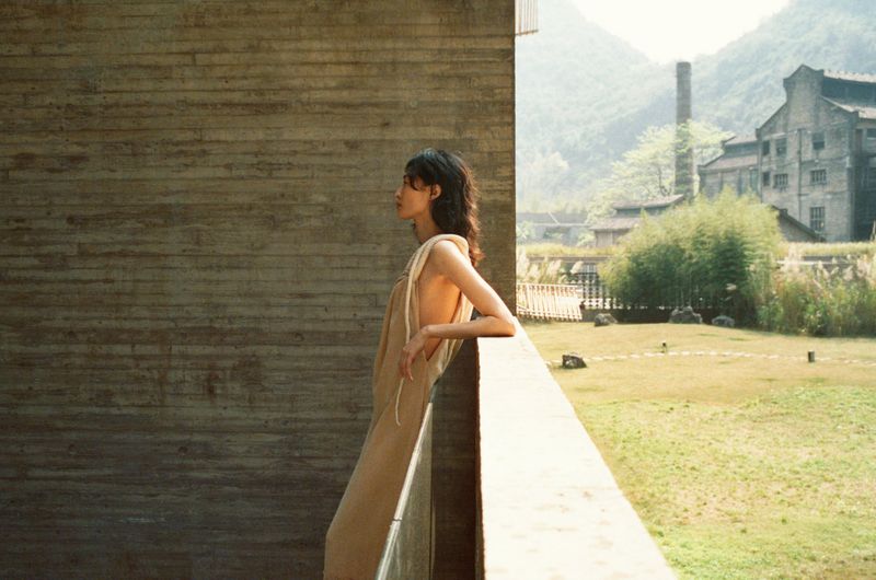 Woman in neutral dress stands on a balcony against mountain backdrop. Minimalist fashion shot, neutral colors, natural light, outdoor setting.