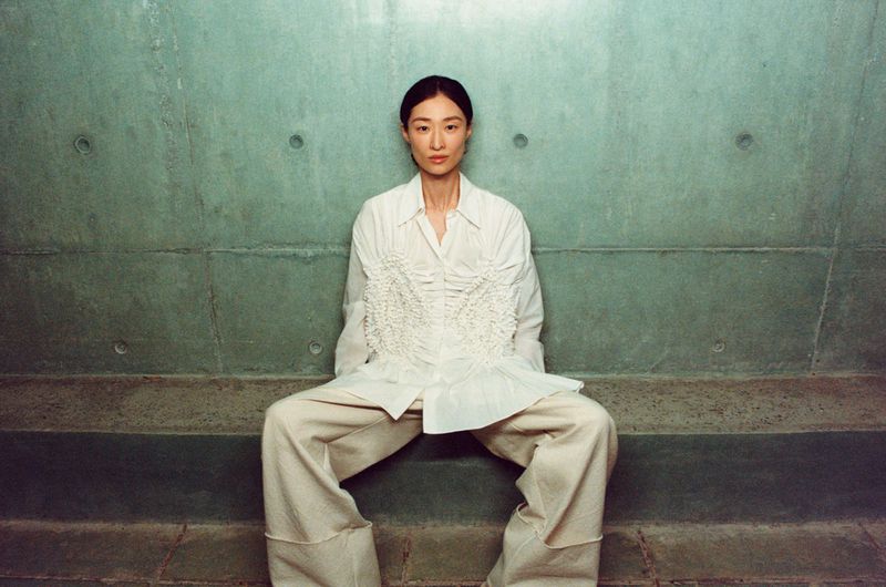 An Asian woman poses in a fashion shot while sitting on a concrete bench, wearing a white shirt and pants against a textured green wall.