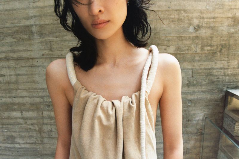 Close-up shot of an Asian woman wearing a neutral dress with rope straps, set against a textured concrete wall backdrop.