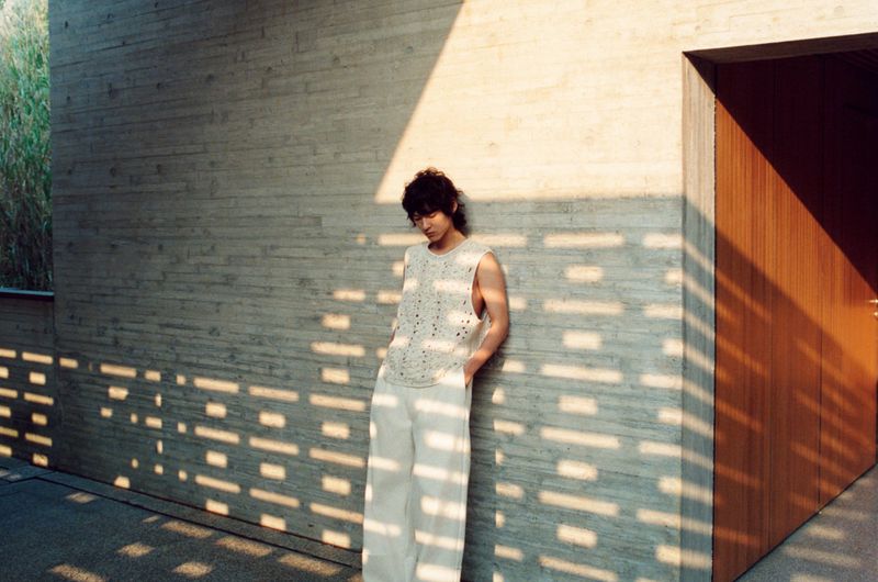 Adult male in a sleeveless shirt and white pants leans against a concrete wall, dappled with sunlight streaming through the reeds outside.