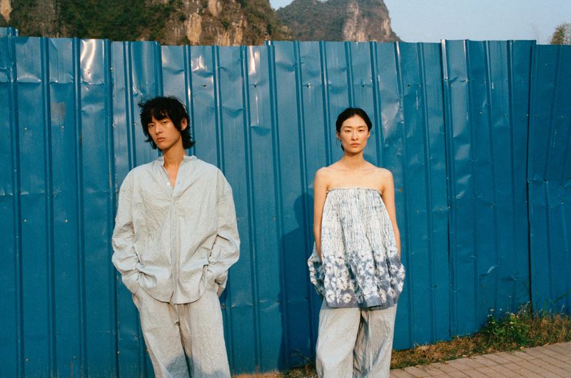 Two Asian adults pose in matching casual outfits against a blue corrugated metal wall with mountains visible in the distance, in an outdoor setting.