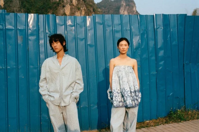 Two Asian adults pose in matching casual outfits against a blue corrugated metal wall with mountains visible in the distance, in an outdoor setting.