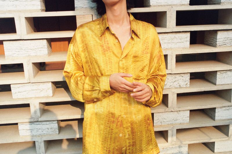 Person wearing yellow reptile-print button-down shirt standing in front of a textured wall during daytime, medium shot.