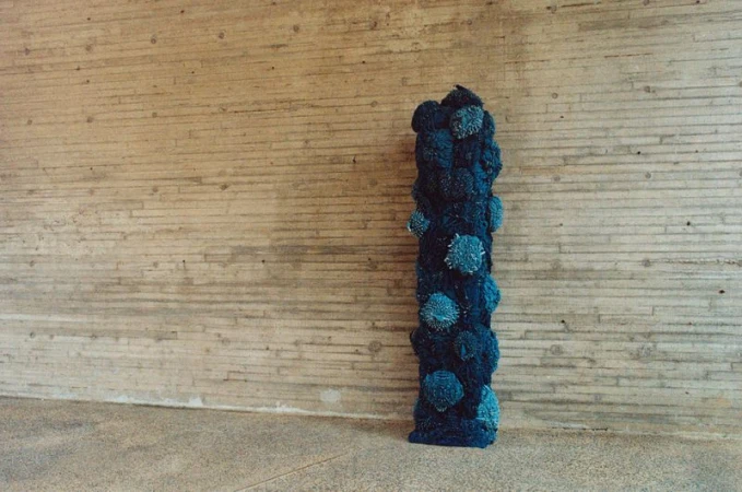 A blue yarn-covered art installation stands against a beige concrete wall, casting shadows on the floor in an indoor setting.