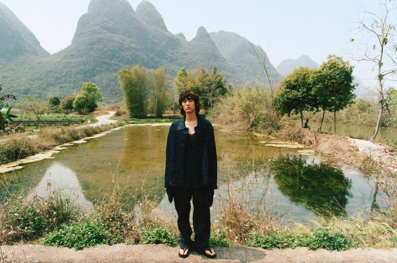 Adult stands near a pond in front of mountains, wearing a dark jacket and pants, captured in a medium shot in natural light.