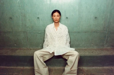 Model seated wearing a white textured blouse and pale trousers against a weathered green concrete wall.