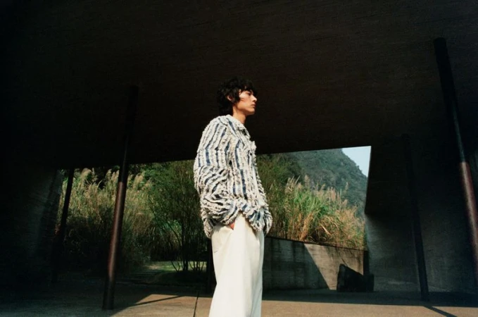 Model wearing a textured patterned jacket and light trousers standing under a concrete structure outdoors