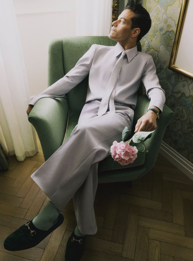 Rami Malek sits in a green armchair holding a pink hydrangea, wearing a light suit and dark loafers in an indoor setting.