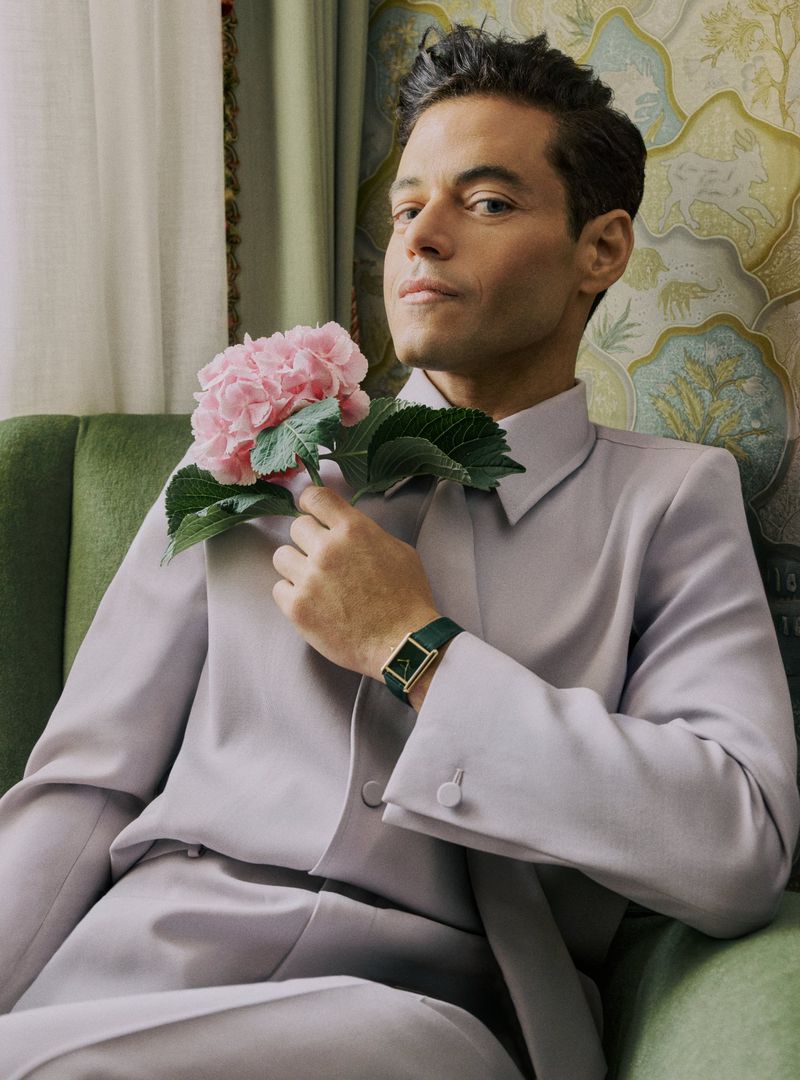 Portrait of a man in a light purple suit holding a pink hydrangea flower while seated on a green chair, posing in an indoor setting.