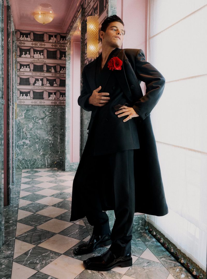 Stylish adult man in a black suit and red flower poses in a pink marble hallway with a checkered floor and decorative wallpaper.