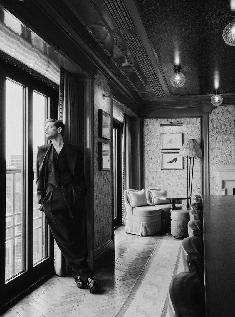 Black and white portrait of an adult man in a suit leaning against a window in an ornate room with a lamp and artwork.