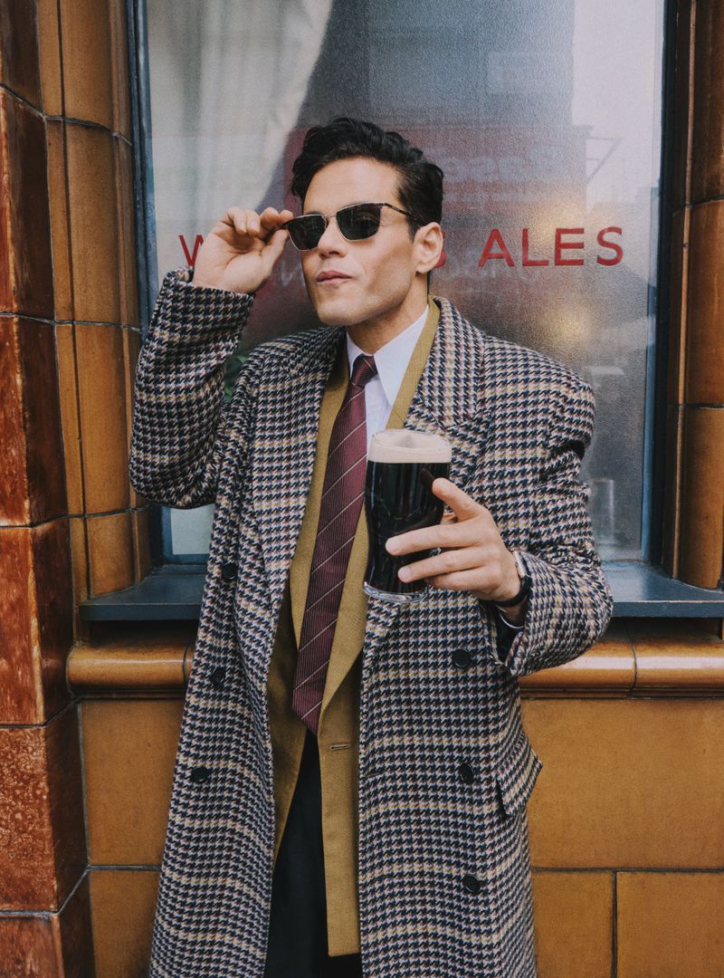 A man in a patterned coat and sunglasses poses while holding a beer next to a vintage building with red text and brown tiling.