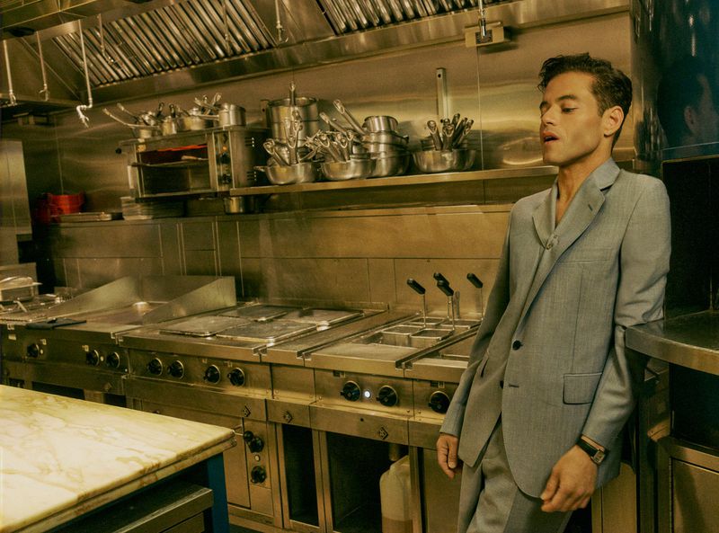 Adult male in a grey suit poses in a commercial kitchen featuring stainless steel appliances and cookware, with neutral, desaturated tones.