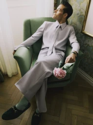 Actor Rami Malek wearing a lilac suit seated in a green armchair holding a pink flower, photographed by Nick Thompson for Dujour.