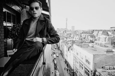 Actor Rami Malek wearing sunglasses, a dark coat, and sweater, seated on a balcony railing overlooking a London street in black and white.