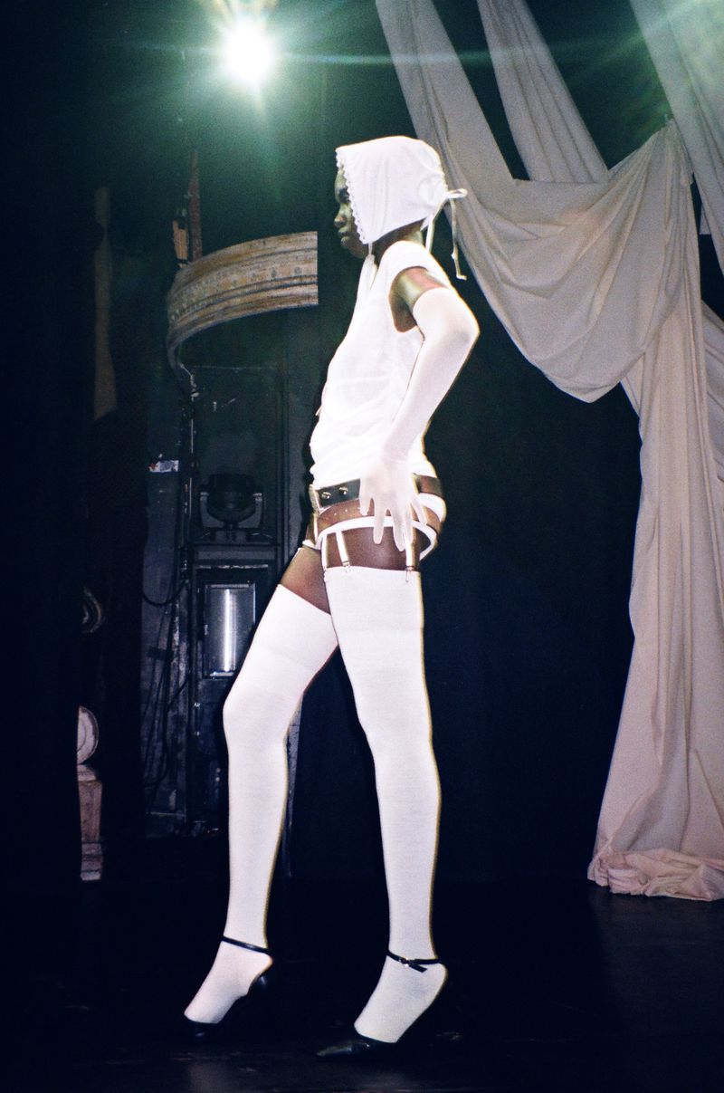 A dark-skinned adult woman in white lingerie and a bonnet poses in a dimly lit room with decorative moldings and sheer fabric drapes.