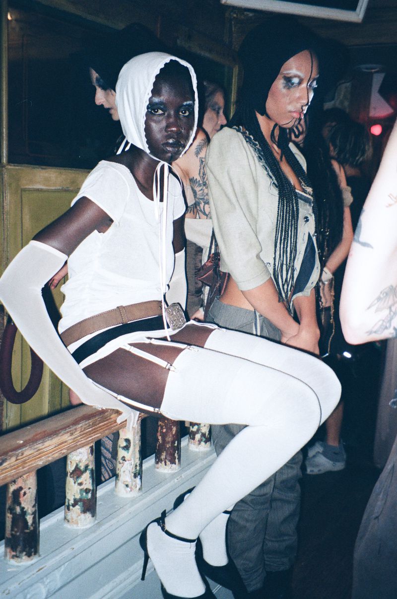 Two women in fashion-forward attire pose indoors near a railing; one is seated and wearing a head covering with strings.