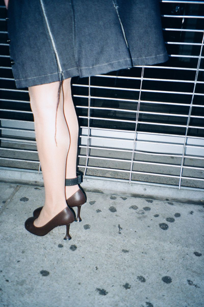 Close-up of a woman's legs in brown heels and a denim skirt standing in front of a metal gate, focus on fashion and urban style.
