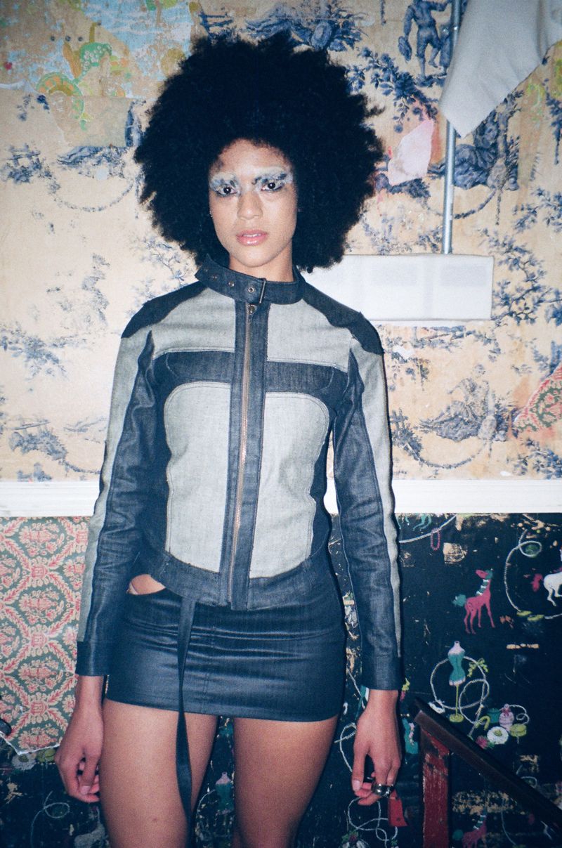 Adult woman with afro wearing denim jacket and skirt in front of floral wallpaper with whimsical drawings, posing indoors.