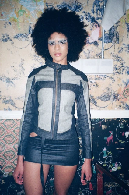 Adult woman with afro wearing denim jacket and skirt in front of floral wallpaper with whimsical drawings, posing indoors.