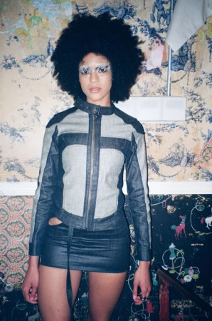 Adult woman with afro wearing denim jacket and skirt in front of floral wallpaper with whimsical drawings, posing indoors.