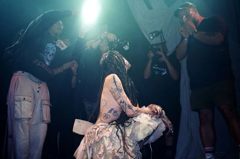 A person with dreadlocks and tattoos in a layered dress at a venue surrounded by people with cameras capturing the scene in a low-lit ambiance.