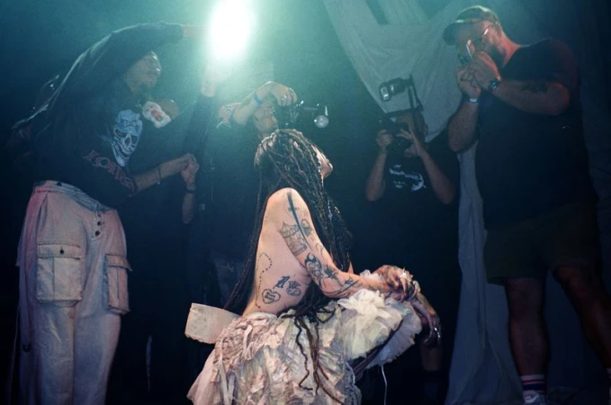 A person with dreadlocks and tattoos in a layered dress at a venue surrounded by people with cameras capturing the scene in a low-lit ambiance.