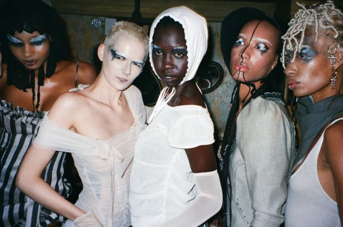 Five models with avant-garde makeup and vintage-inspired dresses pose indoors with neutral lighting, creating a fashion-forward tableau.