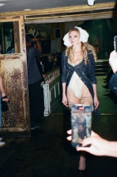 Anna Delvey walking a runway wearing a white ruffled bonnet, an open dark blazer, and a cream bodysuit with garter straps.
