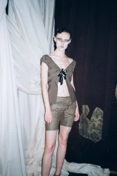 Model wearing olive green top and shorts standing against draped fabric background for Office Magazine Elena Velez project