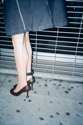 Lower body shot of Anna Delvey wearing a denim skirt, dark brown sculpted high heels, and a black ankle monitor on a concrete sidewalk.