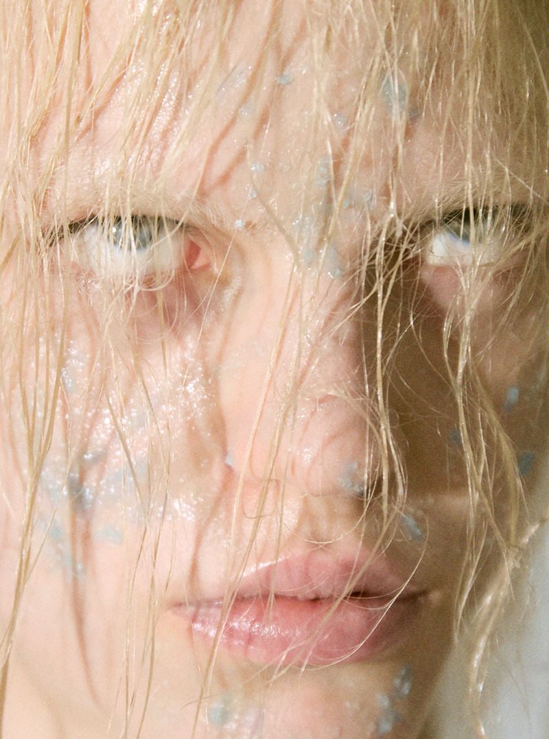 Close-up portrait of an adult woman with wet blond hair and light blue makeup, gazing intently at the camera in a beauty shot.
