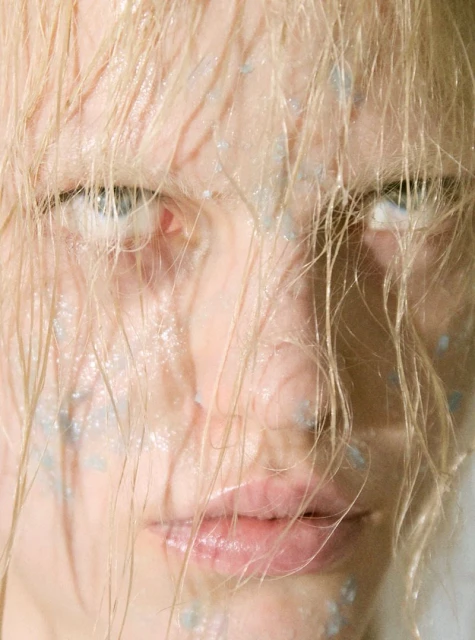 Close-up portrait of an adult woman with wet blond hair and light blue makeup, gazing intently at the camera in a beauty shot.