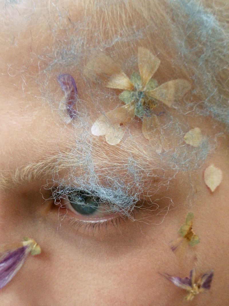 Close-up of a fair-skinned person with blue-tinted light hair, featuring small dried flowers and blue strands on their face.