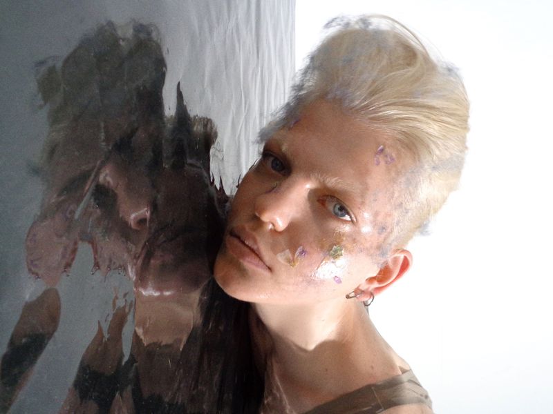 Close-up of a blonde woman with floral accents on her face, posing next to a reflective surface in a brightly lit setting. Beauty shot.