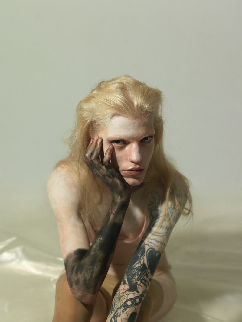 Portrait of a fair-skinned, long-haired person with light hair, arm painted black, posing against a white background, looking at the camera.
