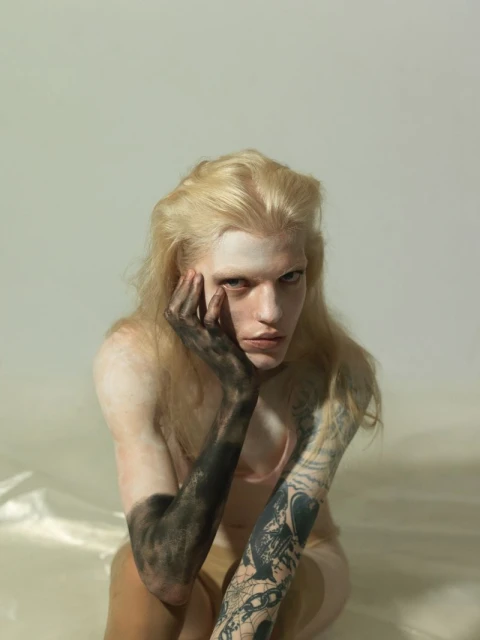Portrait of a fair-skinned, long-haired person with light hair, arm painted black, posing against a white background, looking at the camera.