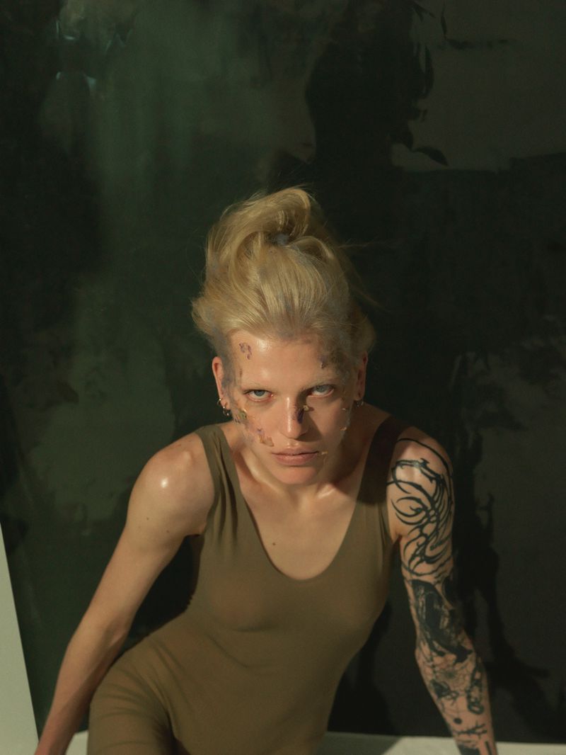 Portrait of a woman with bleached hair and dramatic makeup wearing a dark green tank top against a dark green background.