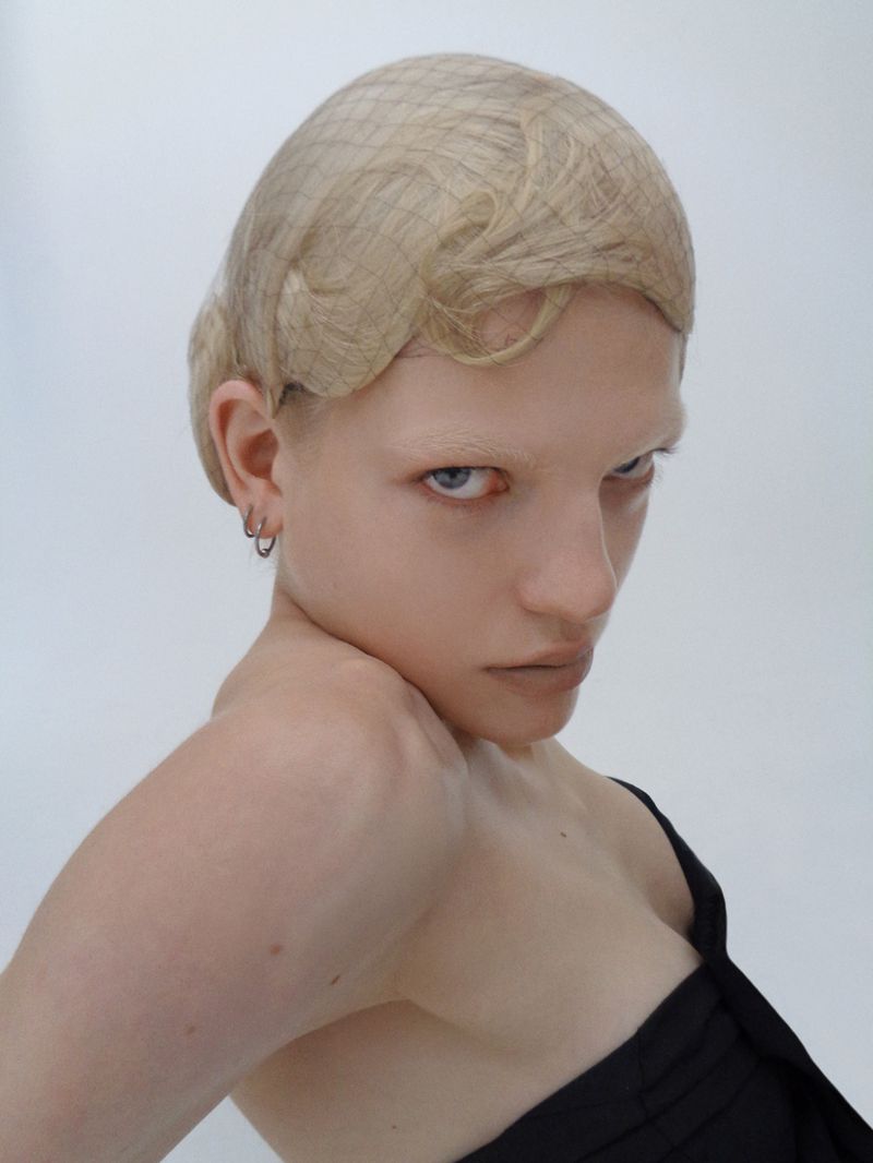 Portrait of a fair-skinned young adult woman with short blonde hair and striking blue eyes, wearing a black top against a white background.