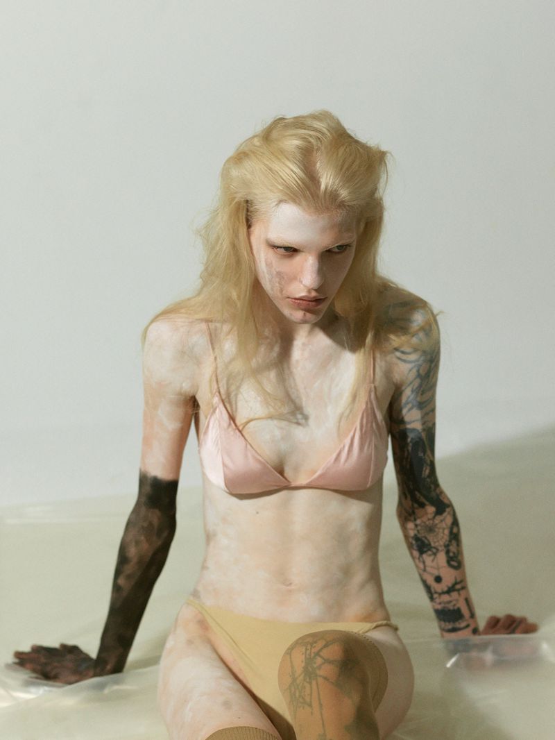 Woman with white hair and partial body makeup in a pink bra and tan bottoms sitting indoors with neutral backdrop.